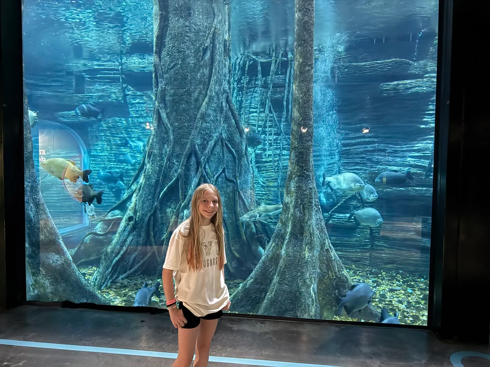 Eliza smiling in front of a huge aquarium tank with big fish swimming among tree roots.