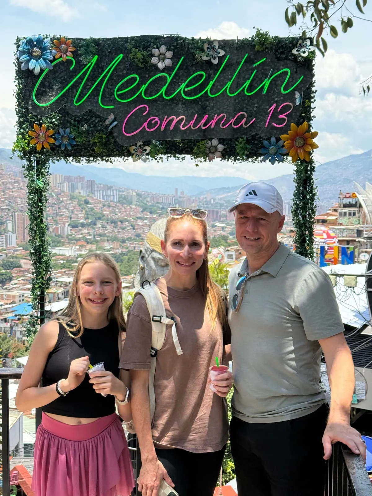 Eliza, Sarah, and me under the neon Medellín Comuna 13 sign, with the hillside neighborhoods and mountains behind us.