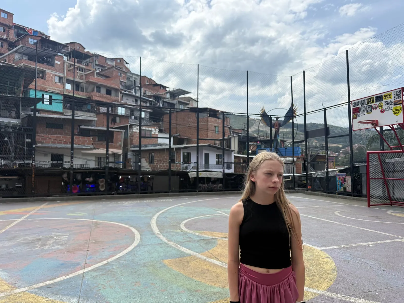 Eliza standing on a brightly painted neighborhood sports court in Comuna 13, with brick homes stacked up the hill behind her.
