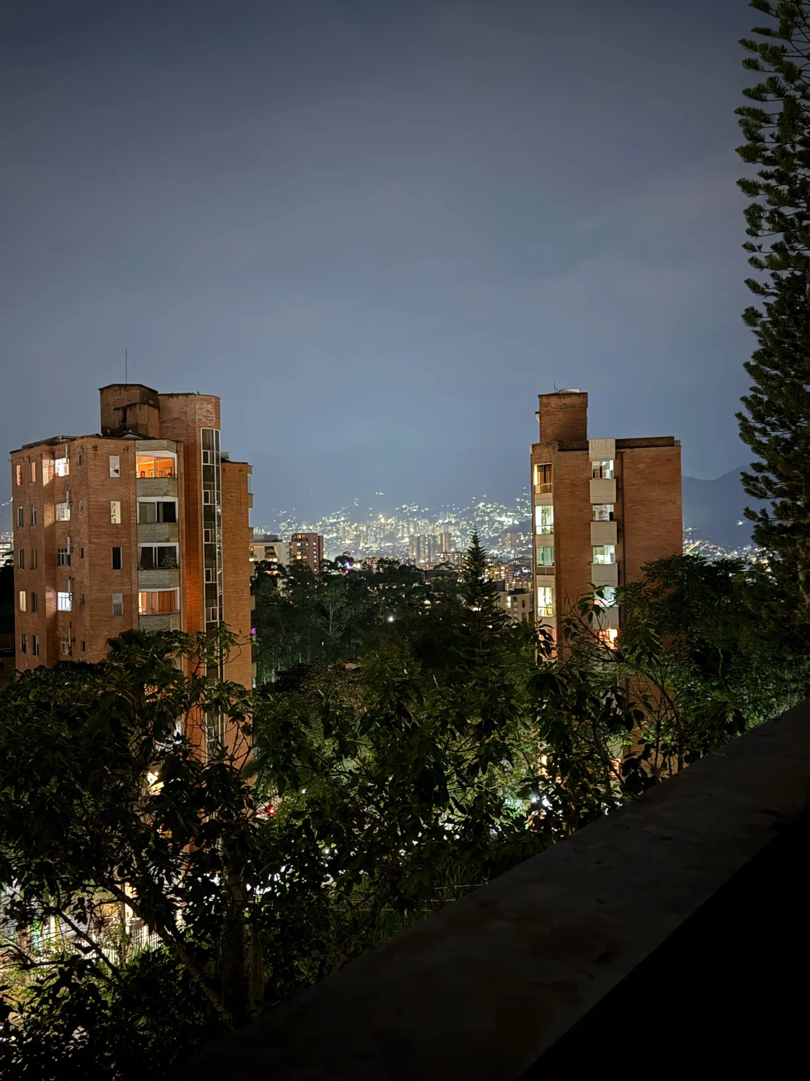 The city lights of Medellín at night, seen between two brick apartment towers and a line of trees.