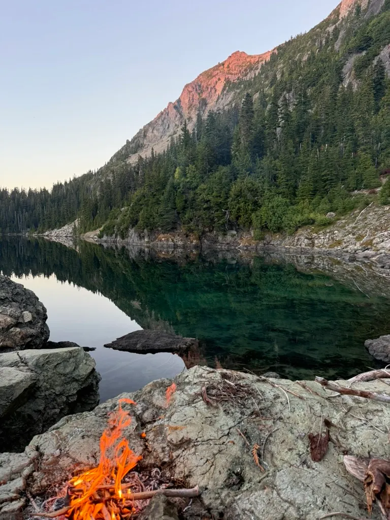 A small campfire on a rock at the edge of a glassy green lake, with the last pink light of sunset on the peak above.