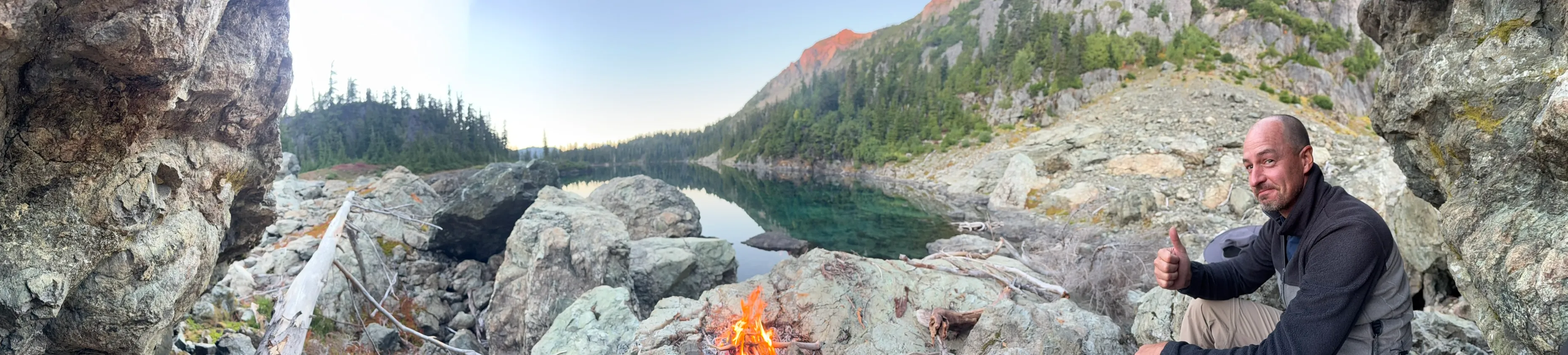 A panorama from a rocky nook above the lake at dusk, with a small campfire in the middle and a thumbs-up on the right.