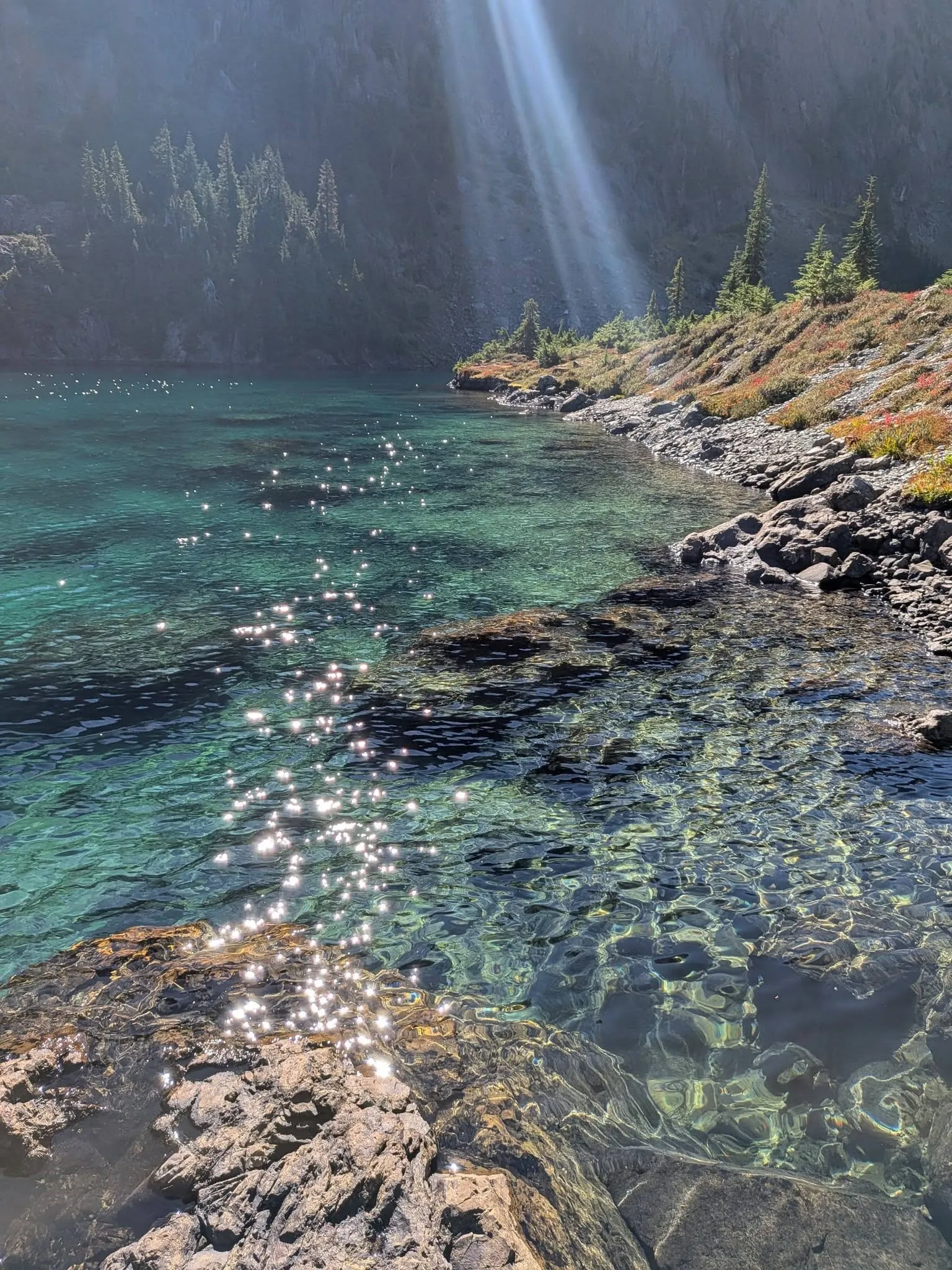 Sunlight streaming down a cliff onto water so clear you can see every rock along the shoreline, with red and gold fall plants on the bank.