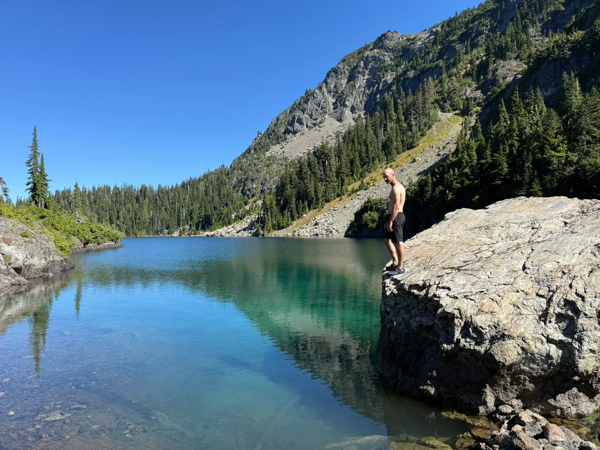 Standing shirtless at the edge of a big boulder, looking down into clear turquoise water, with a rocky ridge and firs across the lake.