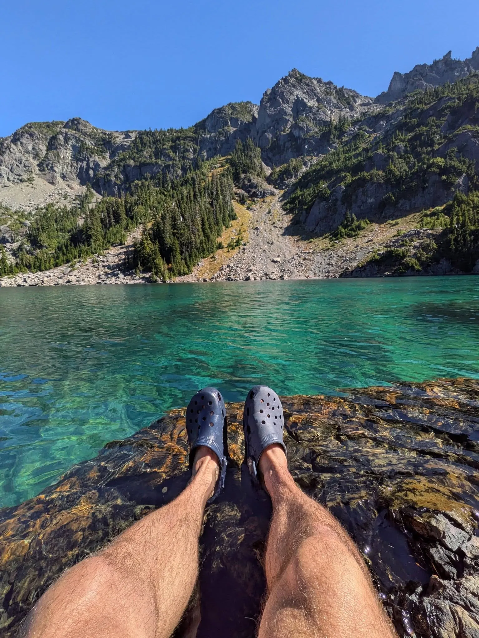 Feet up in blue camp shoes on a rock at the edge of a turquoise lake, looking out at a ring of rocky peaks under a blue sky.