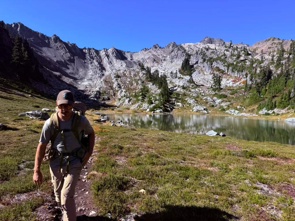 A hiker with a backpack walking up a trail through a green alpine meadow, with a still lake and a wall of pale rocky peaks behind him under a deep blue sky.