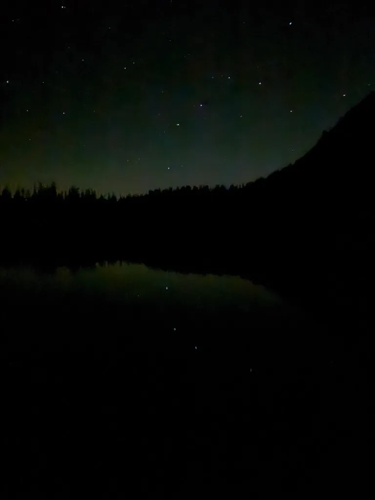 Stars over a dark lake, with a line of trees and a mountain in silhouette and a few stars reflected in the water.