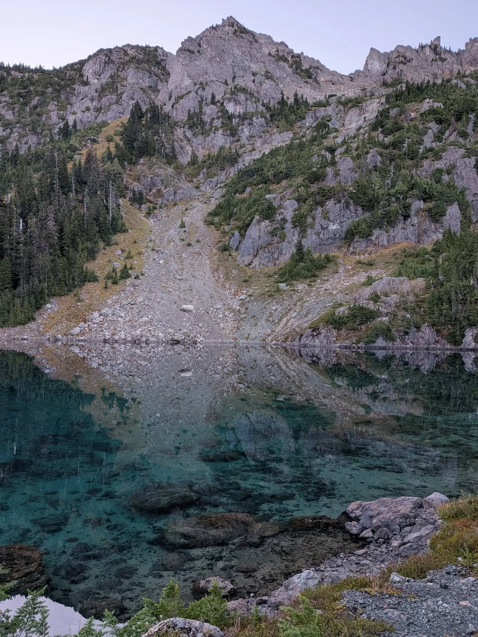 A rocky peak and a long slope of loose rock reflected in still turquoise water, with the lake bottom visible right up to the shore.