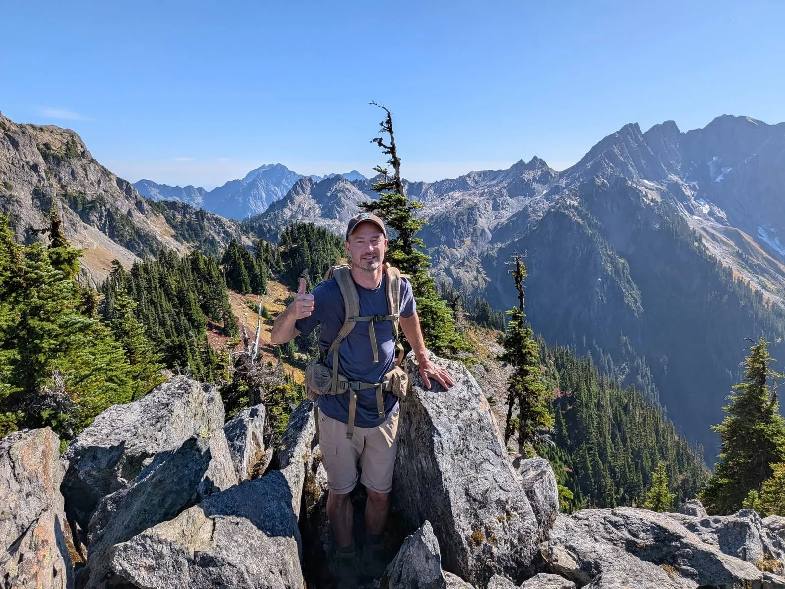 A thumbs-up from a rocky perch above a forested valley, with a long wall of Olympic peaks and a few patches of snow behind.