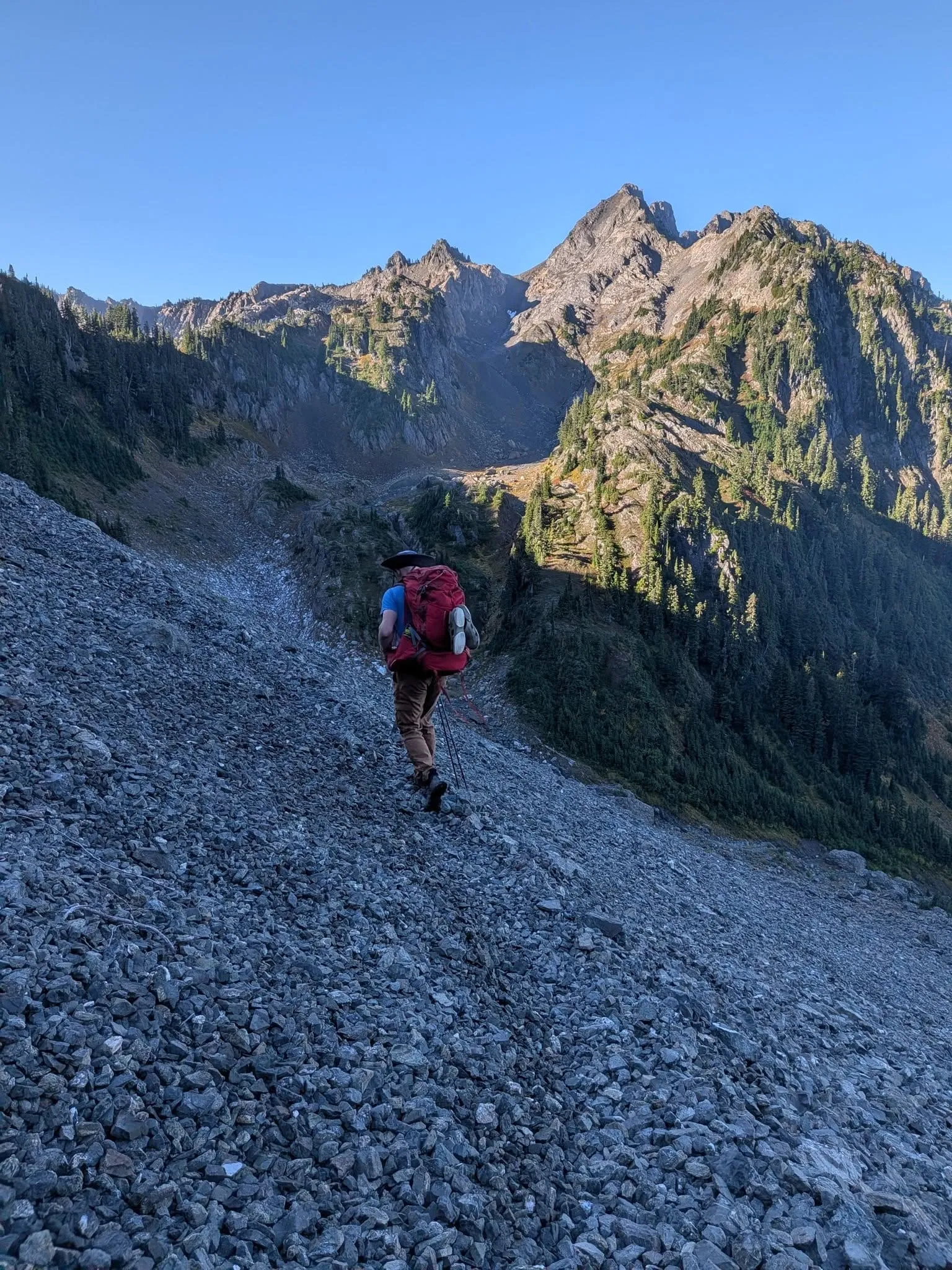 A hiker with a red pack and trekking poles working his way down a steep slope of loose gray rock, with jagged peaks against a clear blue sky.