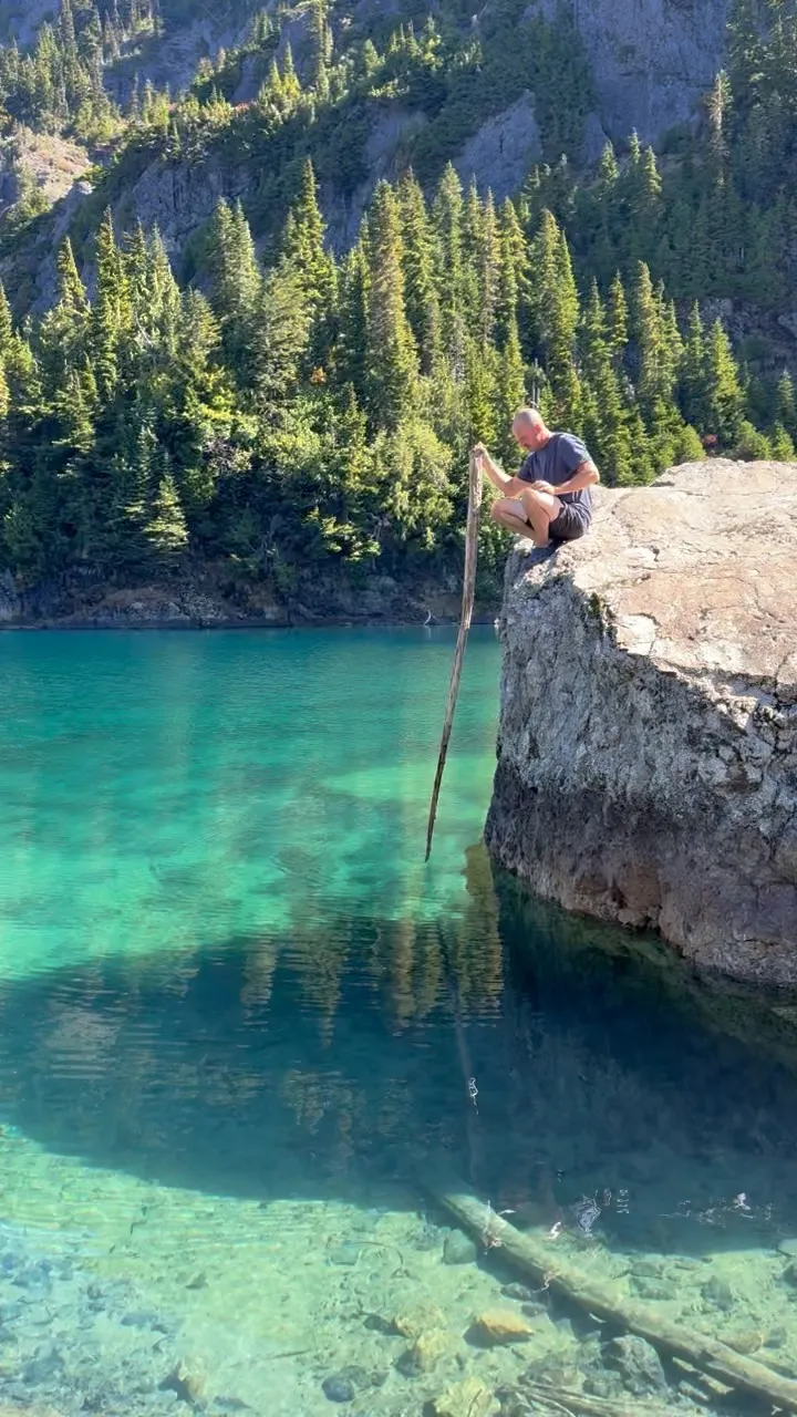 Crouched on a big boulder at the edge of the turquoise lake, holding a long stick that stands straight up out of the water, with the bottom clearly visible below.